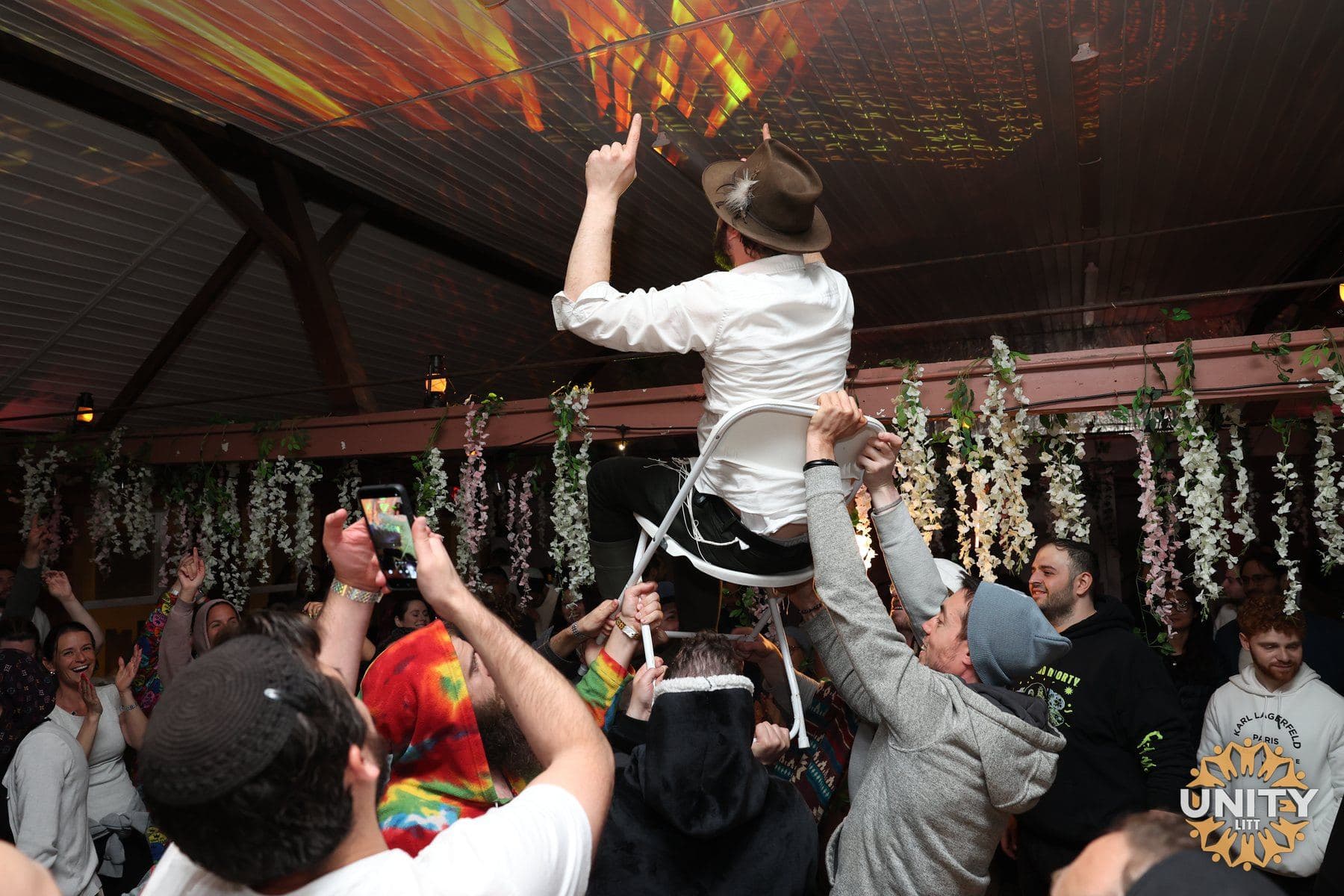 A man lifted on a chair above a dancing crowd, hands reaching up to steady him