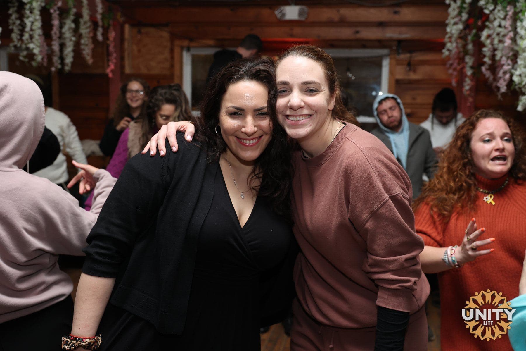 Two friends embracing in the middle of a gathering, both laughing