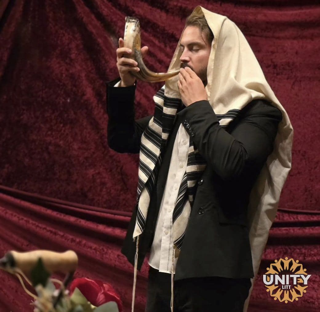 A man wrapped in a tallis blowing the shofar against a deep red curtain