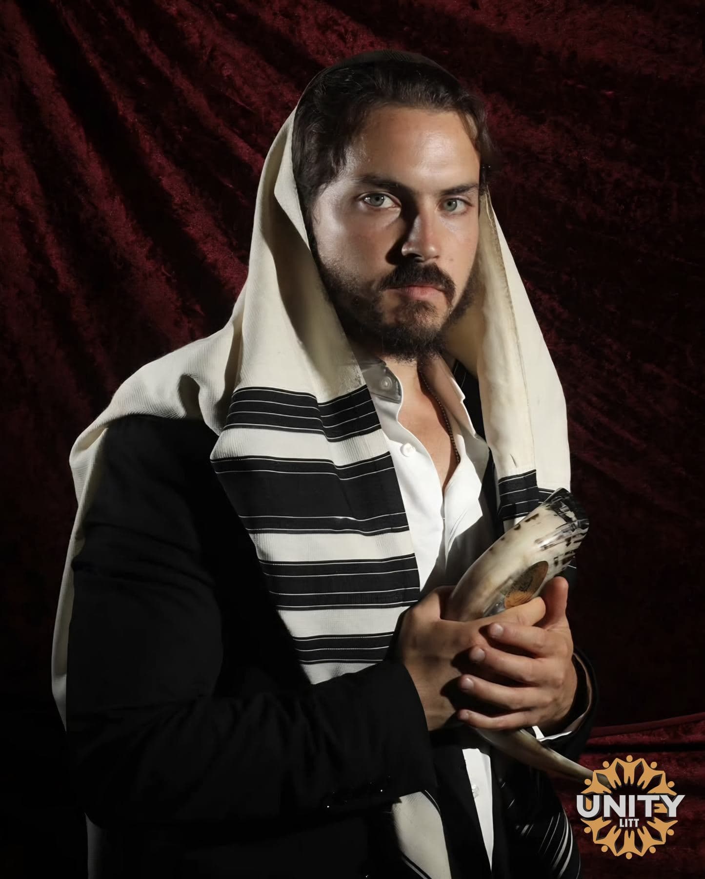 A man in a tallis holding a shofar, photographed against deep red