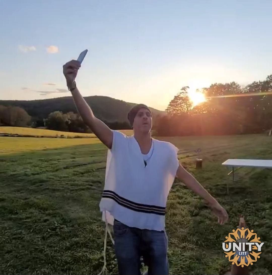 A man in a tallis in an open field at sunrise, one arm raised